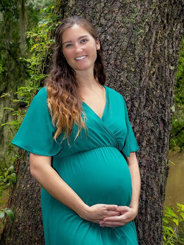 Woman in a green dress propped against a live oak tree cradeling her baby bump. Charleston Maternit
