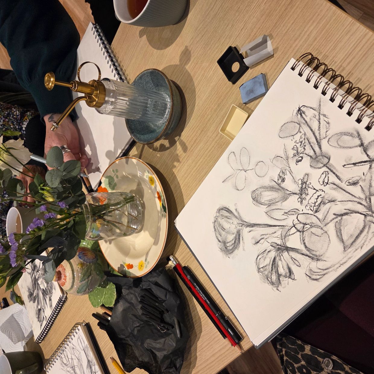 Artists sketch flowers from a vase during a creative drawing session.