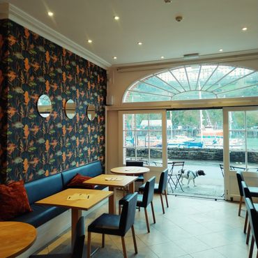 Cozy cafe interior with floral wallpaper, wooden tables, and a dog walking outside near the harbor.