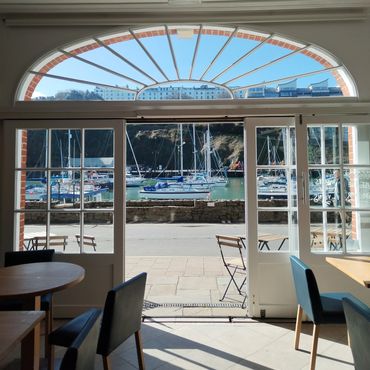 Harbor view from inside a cozy café with tables and chairs near large glass doors.