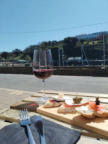 A glass of red wine and appetizers on a wooden board by the waterfront on a sunny day.