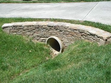 Stone retaining wall with a drainage pipe beneath it on a grassy area.