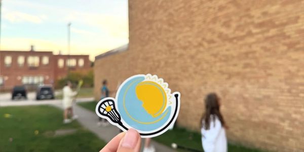 Hand holding a lacrosse-themed sticker with kids playing outside in the background.