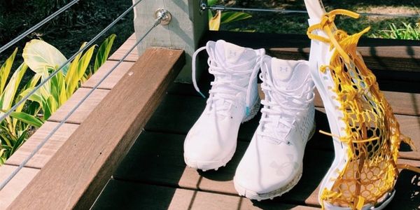 White lacrosse cleats and a lacrosse stick on a wooden deck.