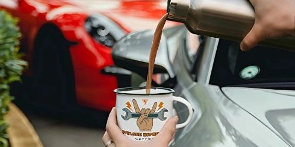 Person pouring coffee into a mug with a wrench logo, near a silver and a red sports car.