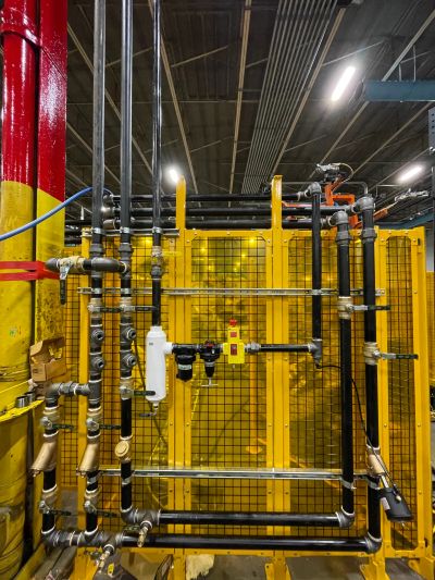Industrial pipes and valves mounted on a yellow grid panel in a factory setting.