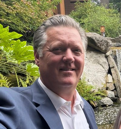 A man in a suit smiles outdoors near a pond and rocks.