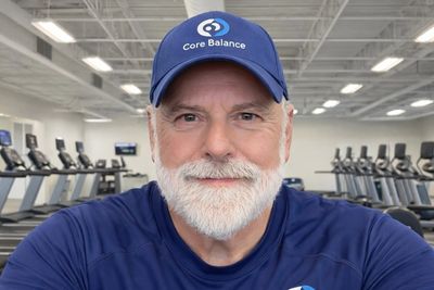 Smiling older man in Core Balance attire at a gym with treadmills.