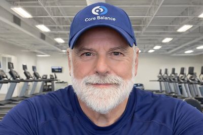 Smiling older man in Core Balance attire at a gym with treadmills.