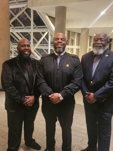 Three well-dressed men posing indoors with smiles.