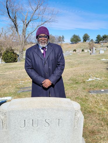 Man in a coat standing by a gravestone engraved with 'JUST' in a cemetery on a clear day.
