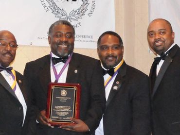 Four men in tuxedos pose with an award plaque at a formal event.
