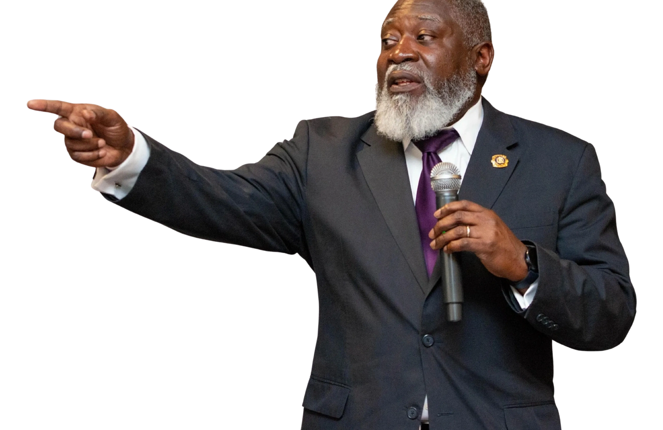 Man in suit with gray beard holding microphone and pointing.