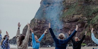 Group practicing yoga on a sandy beach with a rocky cliff backdrop.