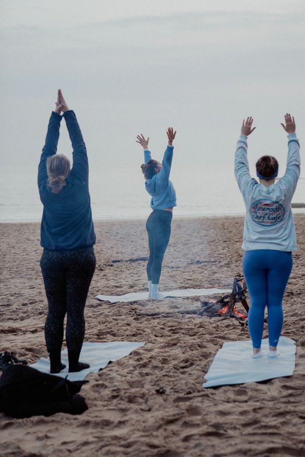 Three people practicing yoga on the beach near a small campfire during early morning.