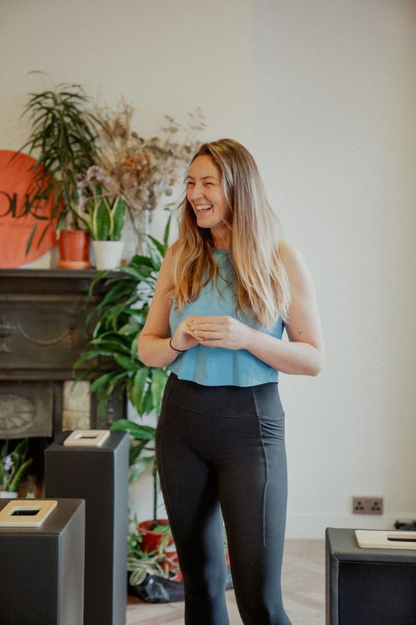 A woman in workout clothes smiling in a cozy room with plants and modern decor.
