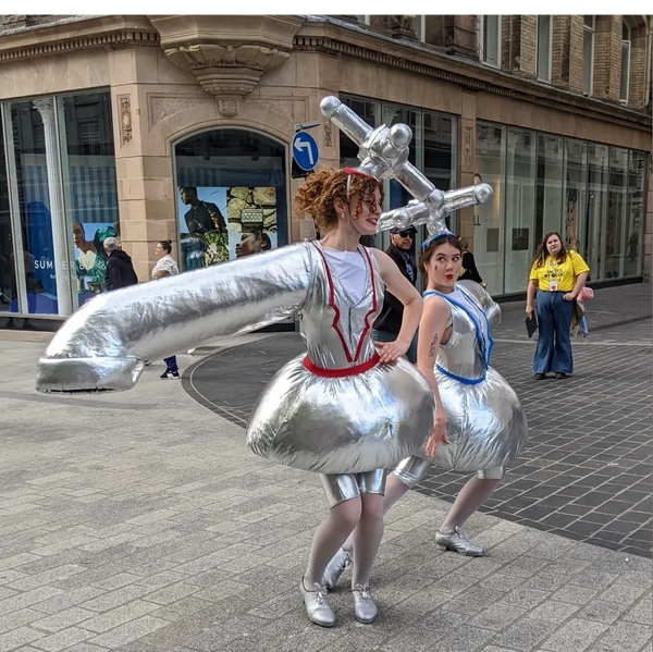 Two women wearing silver costumes like giant bathtaps dance in the street.