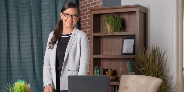 Smiling woman in business attire standing in a cozy office with plants and books.