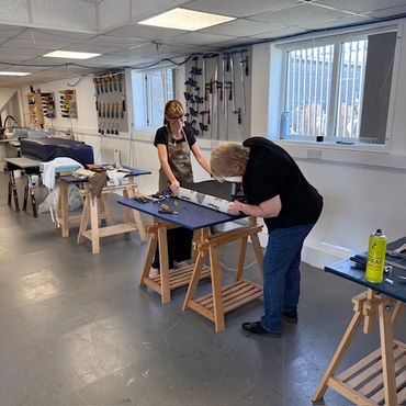 Two women working on a fabric project in a workshop filled with tools and workbenches.