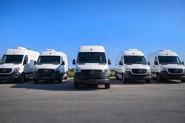 A row of five white Mercedes-Benz vans parked outdoors under a clear blue sky.