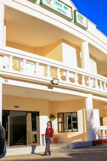 A young girl in a school uniform stands outside Lee's Inn Hotel reception in bright sunlight.