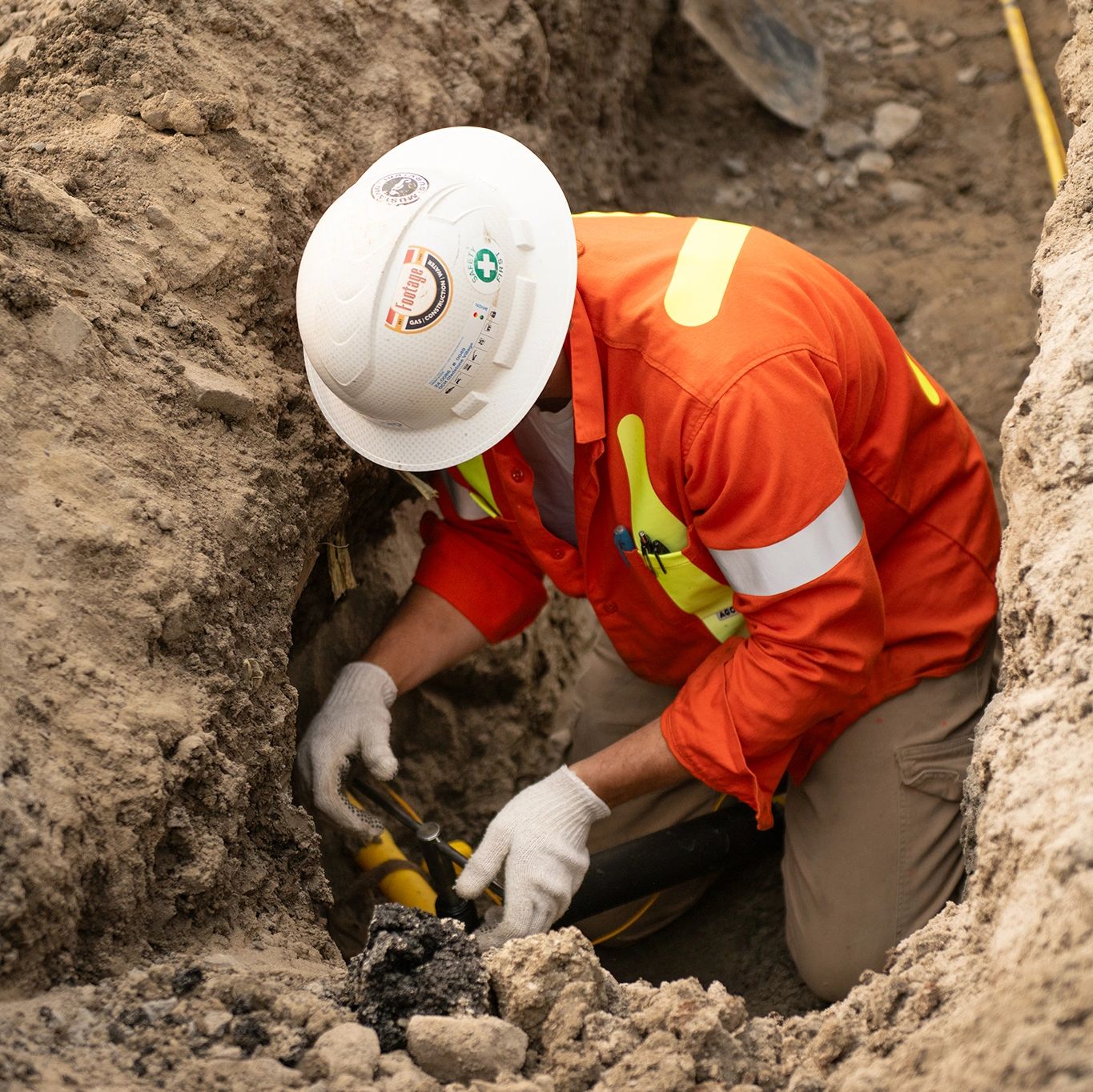 Worker in orange safety gear fixing underground pipes in a trench.