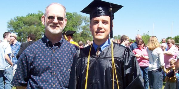 A graduate in cap and gown stands with an older man at an outdoor ceremony.