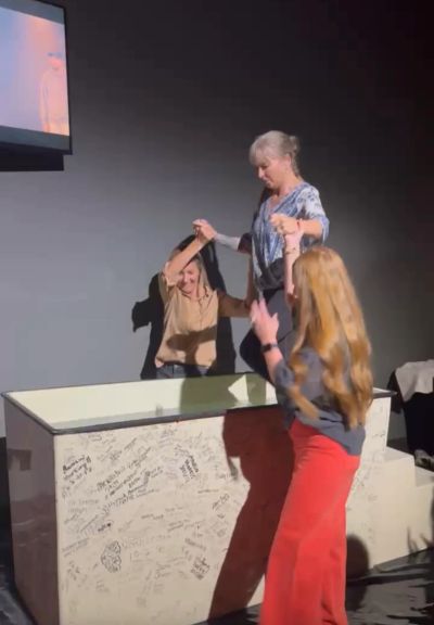 Three women interacting near a signed box in a dark room.