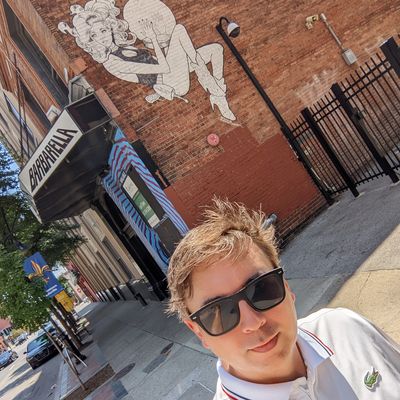 A man wearing sunglasses takes a selfie on a sunny street with a mural behind him.
