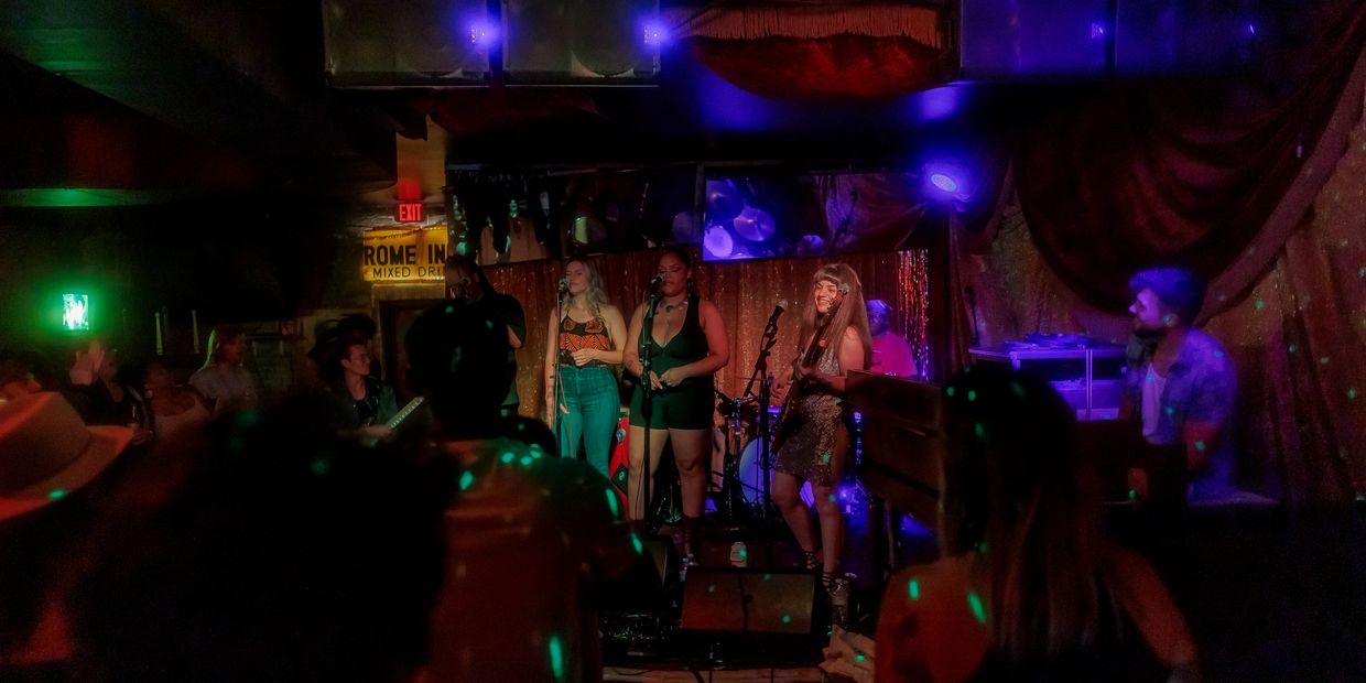 A lively band performs on stage in a dimly lit club with colorful lights.