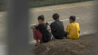 Three boys sitting on a mound by an empty paved area, viewed from behind.
