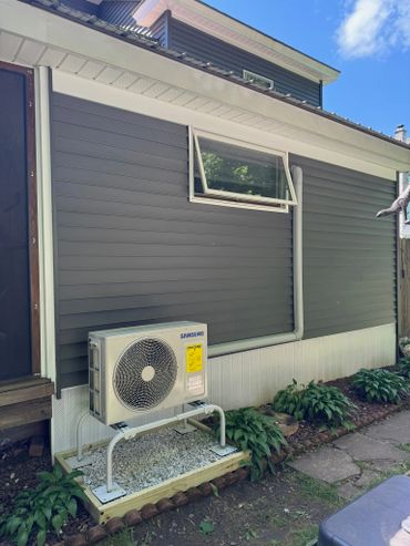 Outdoor Samsung air conditioning unit mounted on gravel bed beside a house.