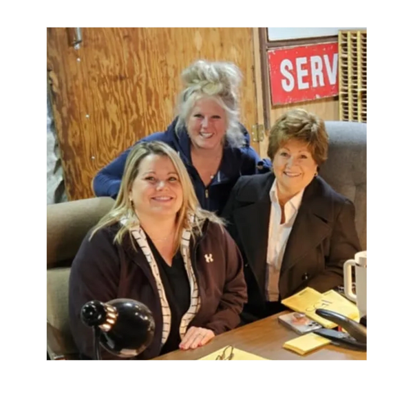 Three women smiling together in an office setting with wooden walls.