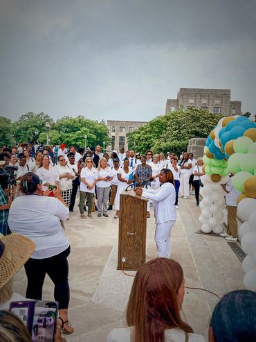 Woman in white speaking at an outdoor podium to a crowd.