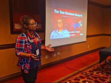 A woman giving a presentation titled The Teen Resilience Blueprint to a small audience.