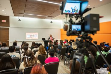 A speaker addresses an audience in a conference room, with a camera recording the event.