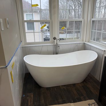 Modern white freestanding bathtub in a corner with large windows and wood-look floor.