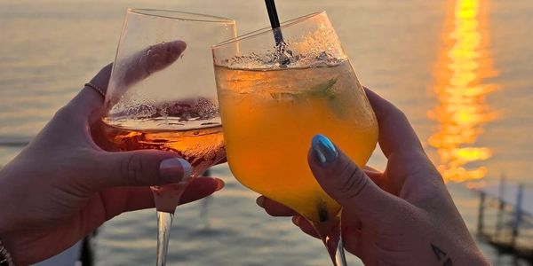 Two hands clinking cocktail glasses at sunset by the water.
