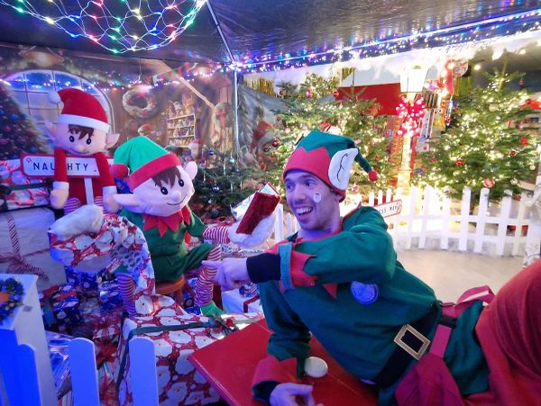 Person dressed as an elf among Christmas decorations and gifts.