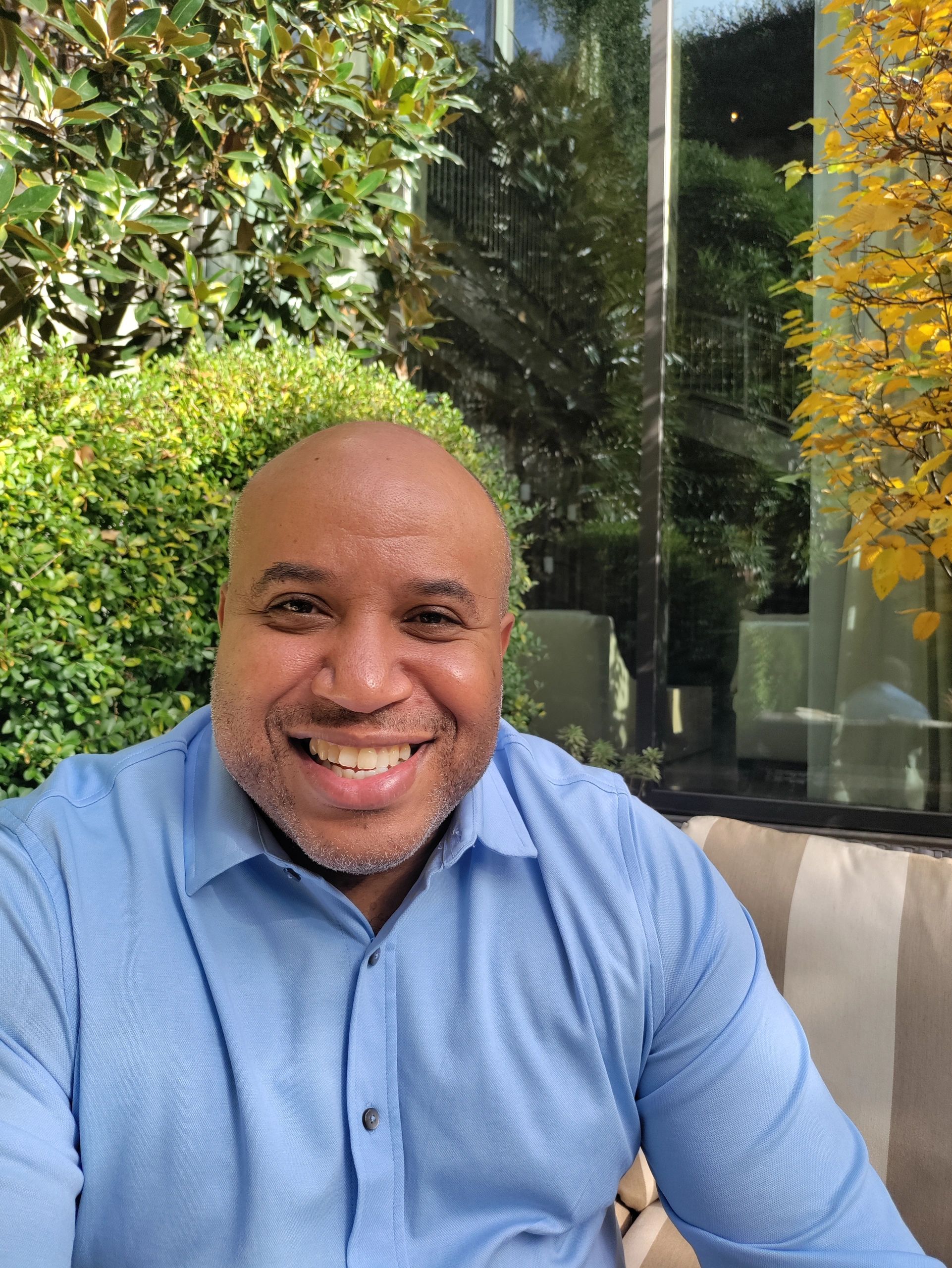 Smiling man in a light blue shirt sitting outdoors with greenery and yellow leaves behind.