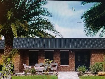 Brick house with black metal roof and outdoor seating area under palm trees.