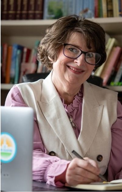 Smiling older woman writing notes at desk with books in background.