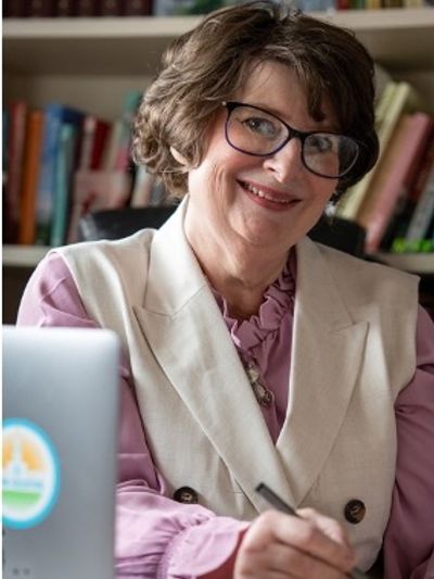 Smiling woman in glasses working at a desk with books in the background.