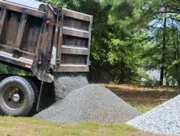 Dump truck unloading gravel on a grassy area near trees.