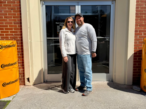 A smiling couple stands outside a building entrance on a sunny day.