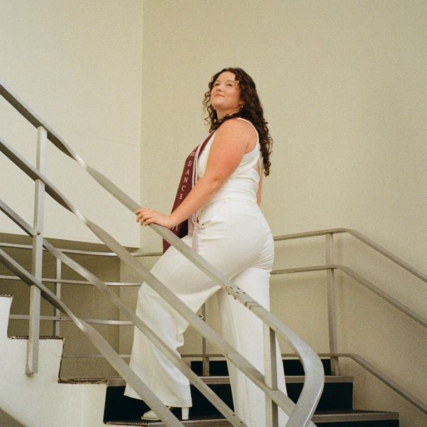 Confident graduate in white outfit posing on staircase with maroon sash.
