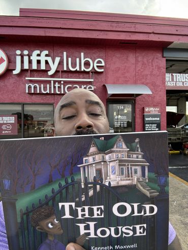Man holding a book titled 'The Old House' in front of a Jiffy Lube.