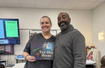Two adults smiling, one holding a book titled 'The Old House' in a classroom.