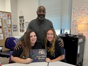 Three people smiling in a classroom, two holding a book titled 'The Old House'.