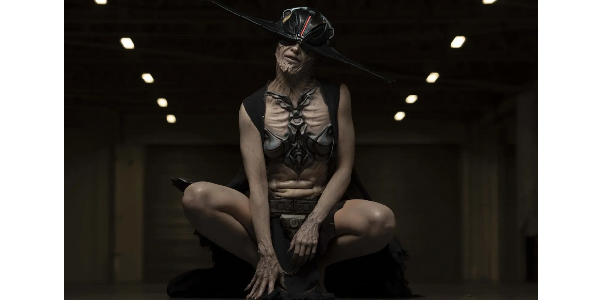 A dark being is crouched on the floor wearing a large prostheic prop